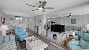 Een moderne woonkamer met een plafondventilator, een flatscreen-televisie en lichtblauwe banken.