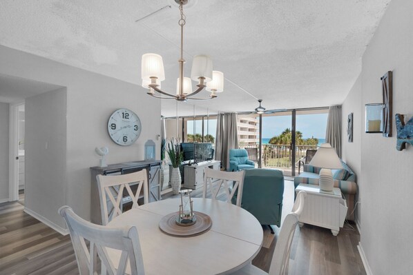Uma sala de estar ampla com uma mesa de jantar redonda, cadeiras brancas, um lustre e uma grande janela com vista para o oceano.