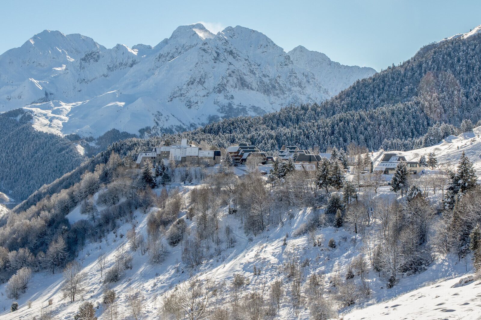 Uma aldeia montanhosa coberta de neve, com edifícios em estilo chalé, rodeada por encostas arborizadas e picos nevados.