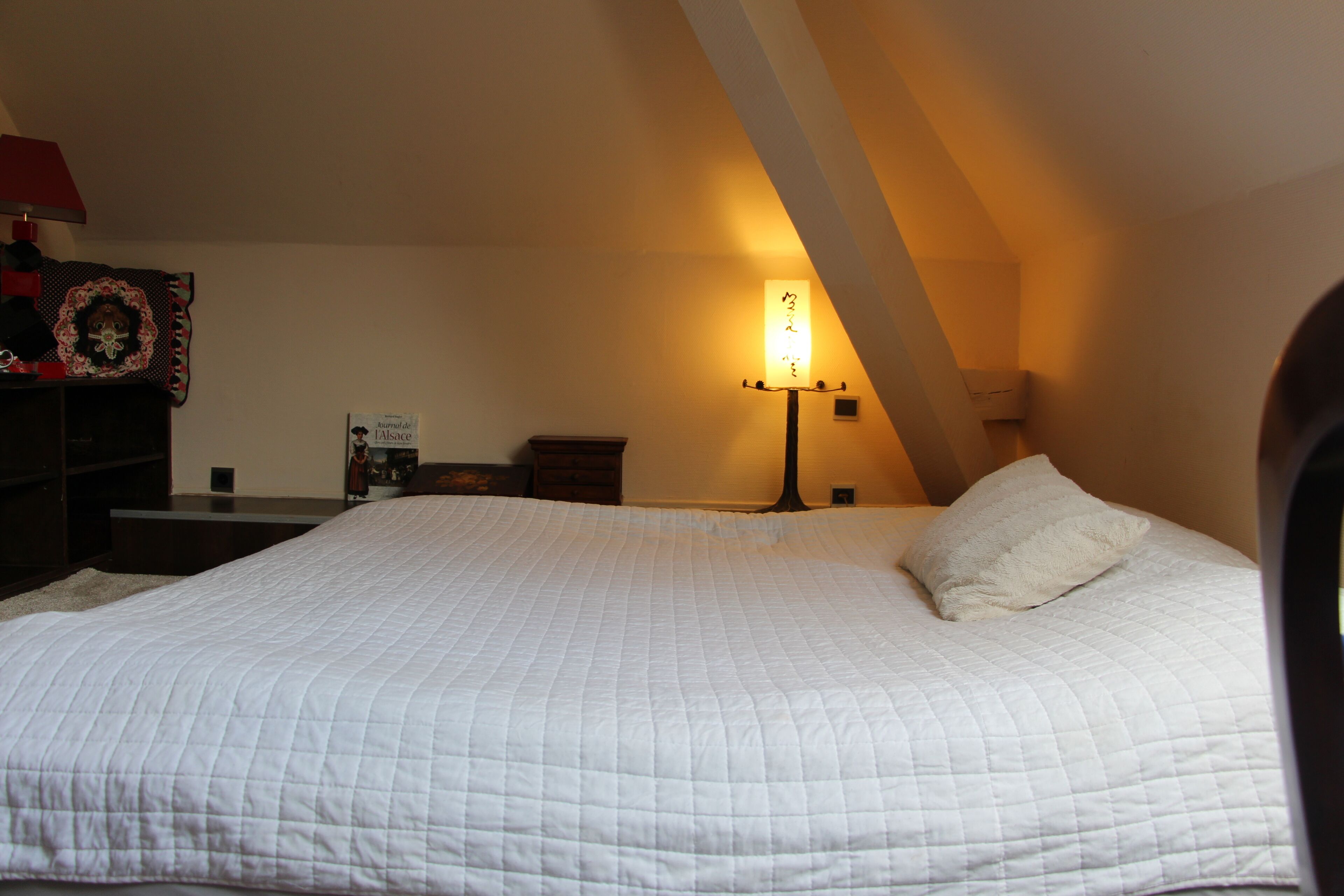 A bedroom with a large bed, a bedside table, a lamp, and a wall-mounted shelf.