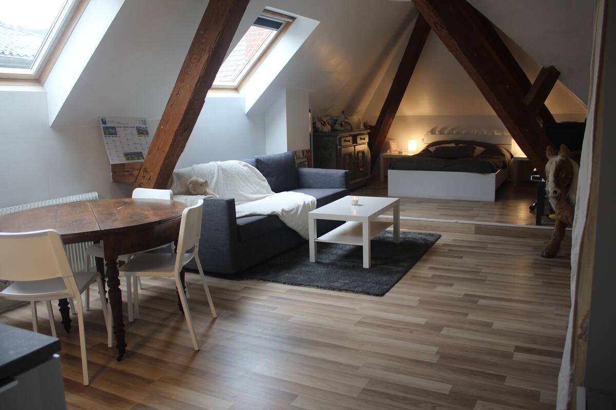 Ein Schlafzimmer im Dachgeschoss mit schrägen Wänden, einem Holzessttisch, einer Couch, einem kleinen weißen Tisch und einem Bett mit Kopfteil.
