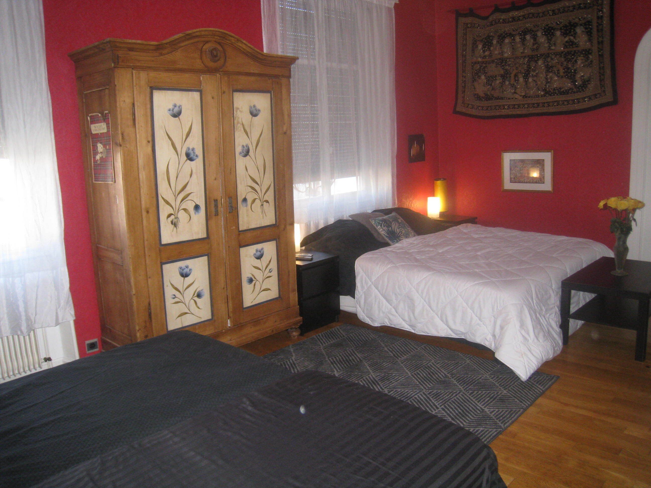 A bedroom with two beds, a wardrobe, and a red wall with a tapestry.
