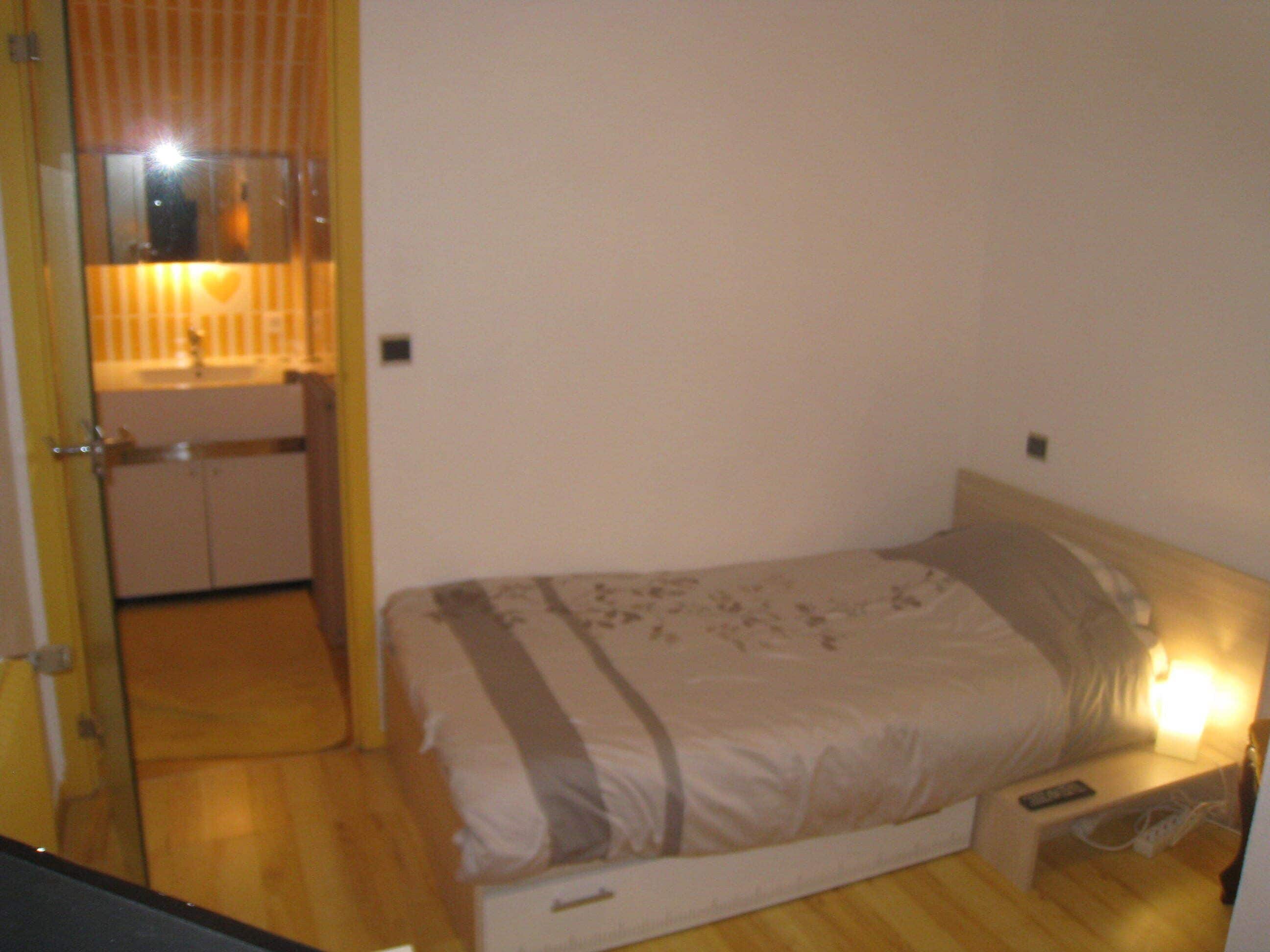 Un dormitorio con cama, mesita de noche, control remoto y baño con espejo y lavabo.