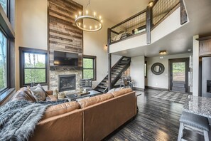 A spacious living room with a large brown leather sofa, a fireplace, a TV mounted on the wall, and a staircase leading to an upper level.