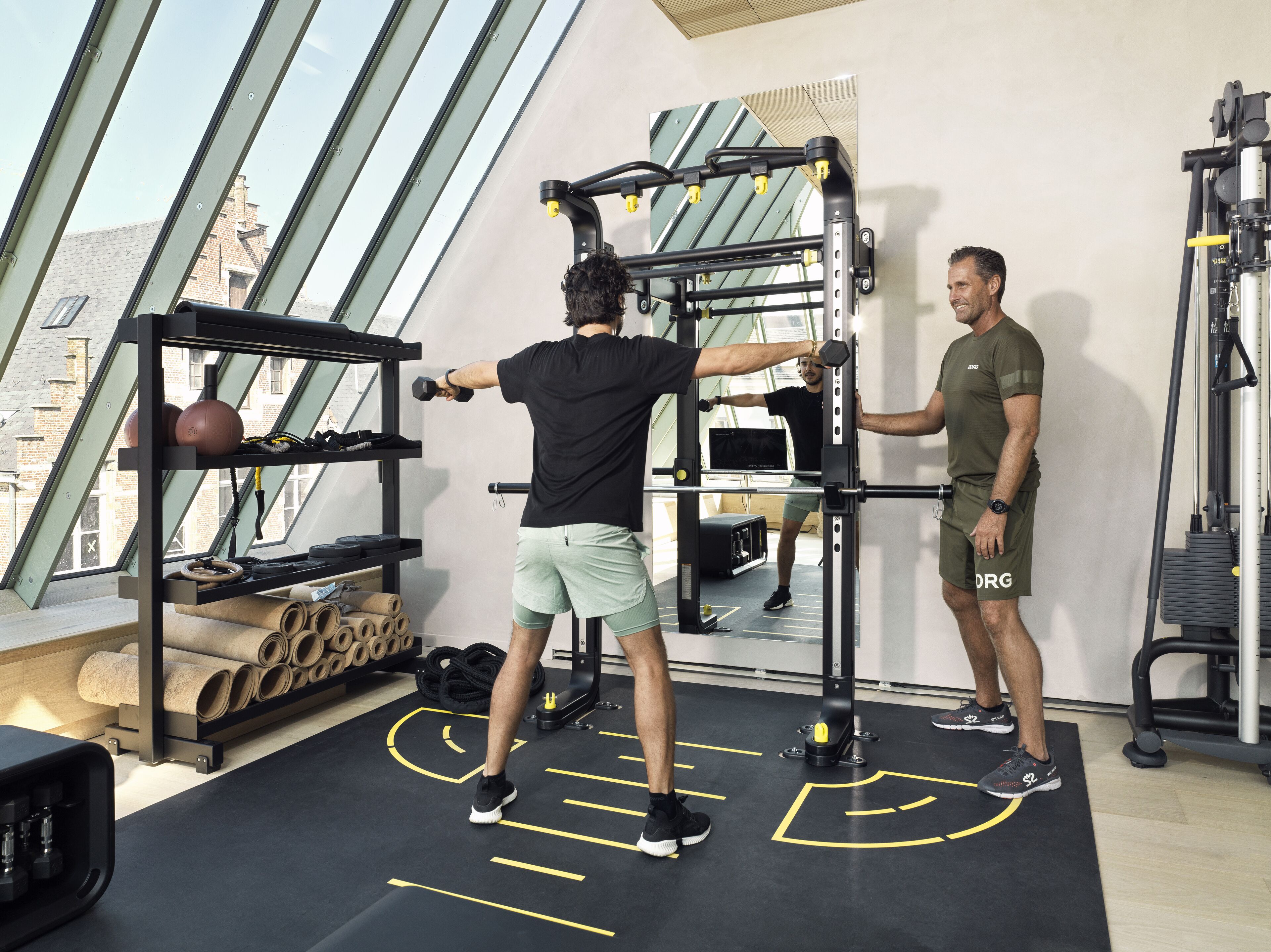 Une salle de sport équipée de matériel d’exercice, d’un miroir et d’une fenêtre donnant sur des immeubles.