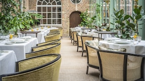 Restaurante com mesas preparadas para refeições, cadeiras de vime e grandes janelas com plantas.