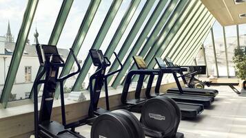 Fila de bicicletas estáticas y cintas de correr en un gimnasio moderno con ventanas amplias y vista a edificios.