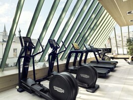 Fila de bicicletas estáticas y cintas de correr en un gimnasio moderno con ventanas amplias y vista a edificios.