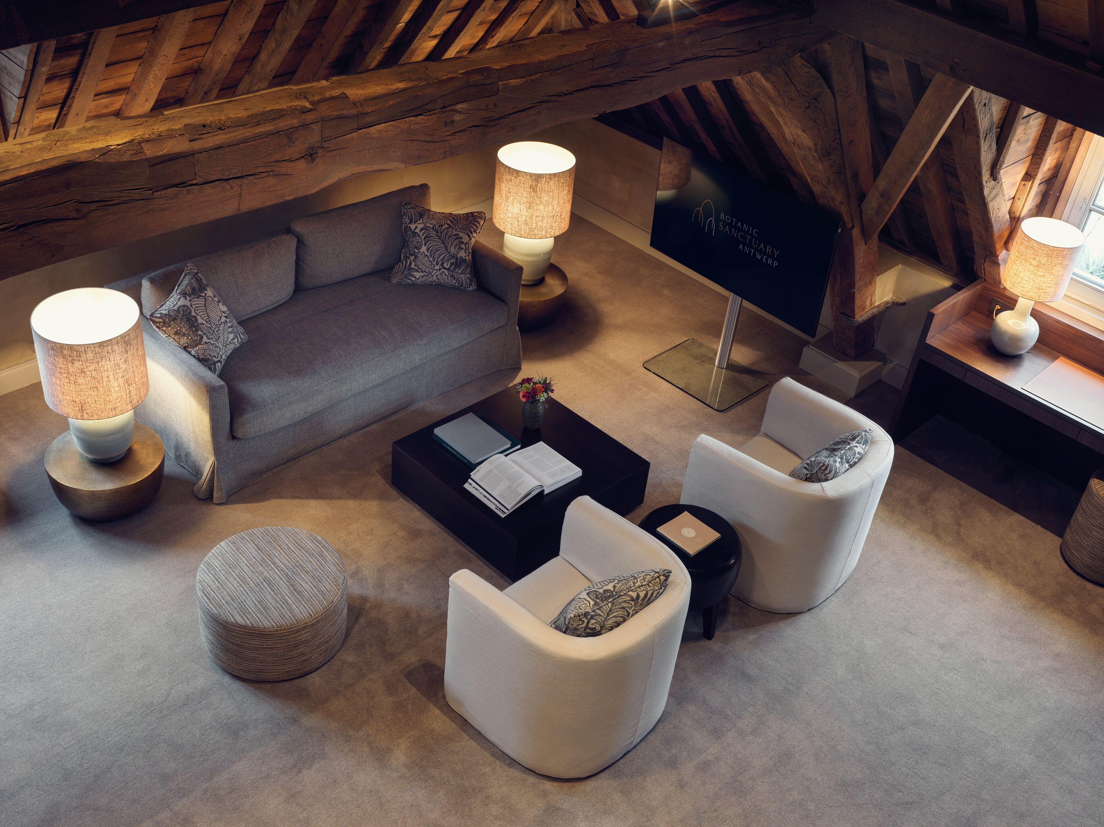 A cozy loft-style living room with a high ceiling, exposed wooden beams, a grey sofa, white armchairs, and a coffee table.