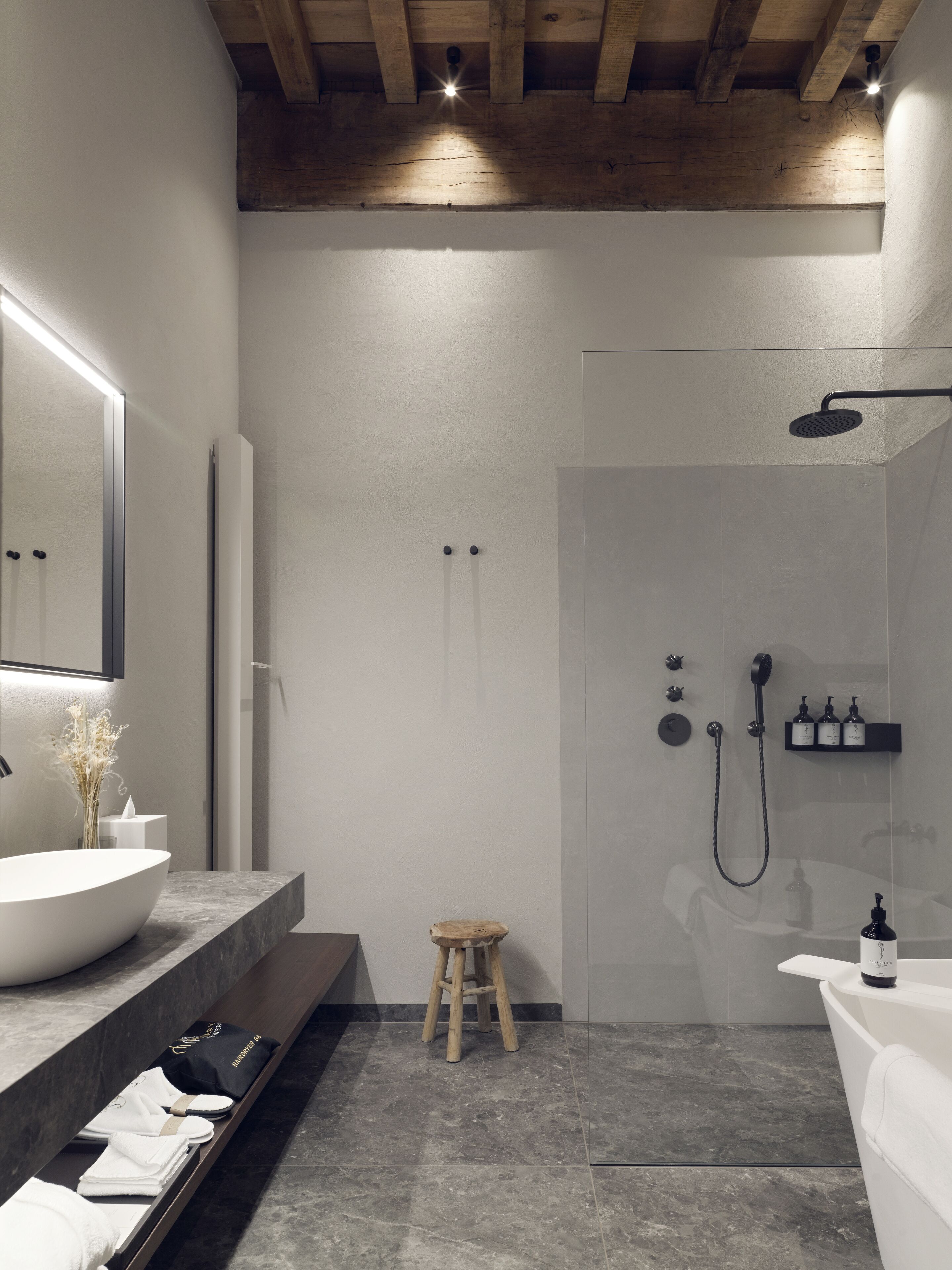 Un baño moderno con un lavamanos grande de color blanco, un taburete de madera, una cabina de ducha de cristal y un estante empotrado con artículos de aseo.