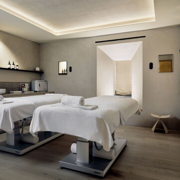 A modern spa room with two massage tables, a large mirror, a sink, and a shelf with toiletries.