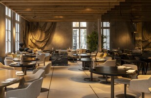 A modern restaurant with round tables, comfortable seating, and large windows allowing natural light.