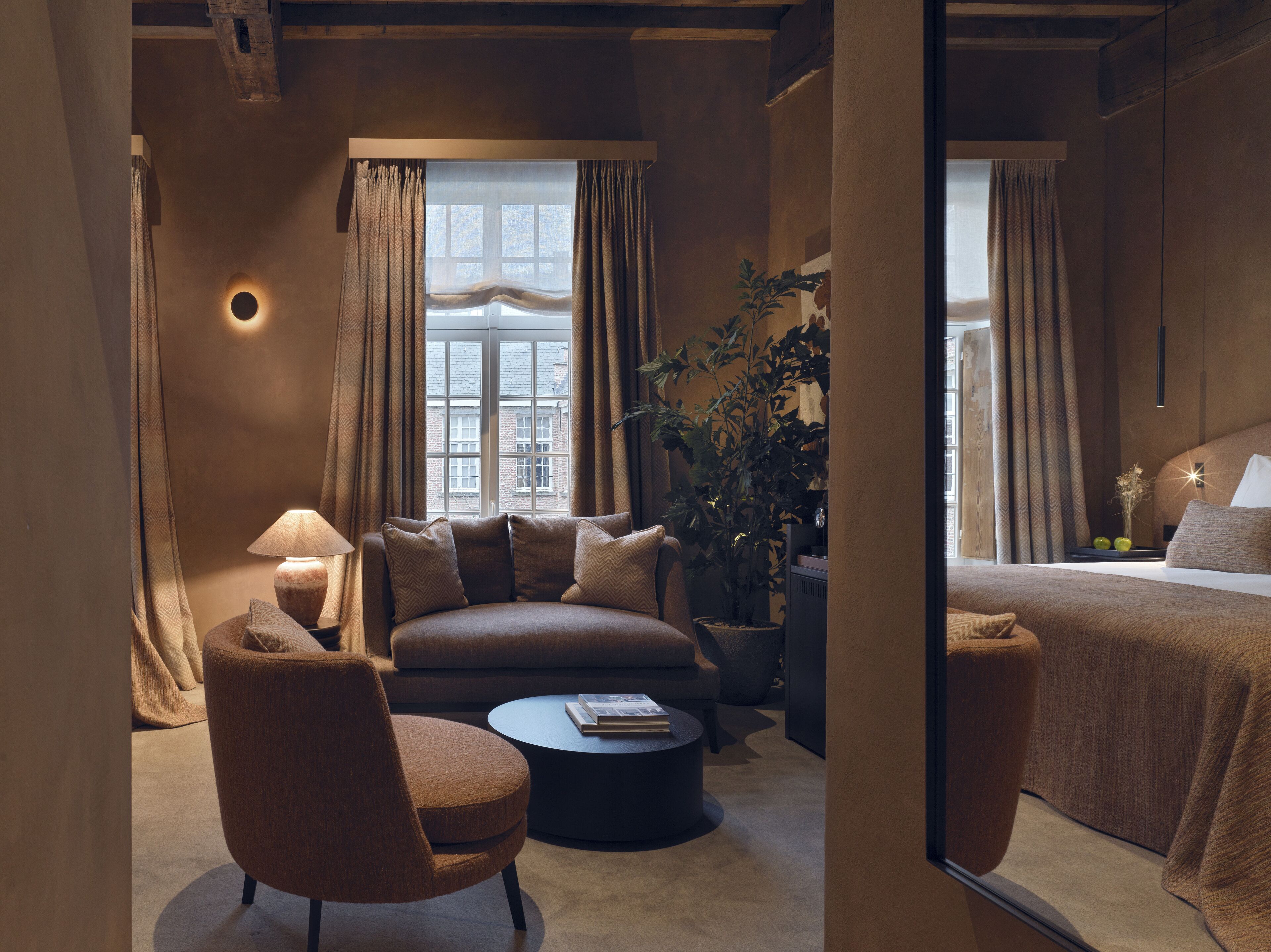 A cozy hotel room with a sofa, armchair, and a small round table. There is a large window with curtains, a potted plant, and a bed with a headboard.