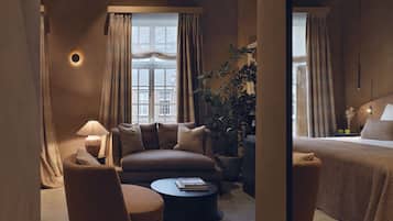 A cozy hotel room with a sofa, armchair, and a small round table. There is a large window with curtains, a potted plant, and a bed with a headboard.