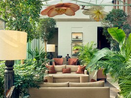 A cozy indoor seating area with a sofa, a table, and a lamp, surrounded by lush green plants and decorative flowers.