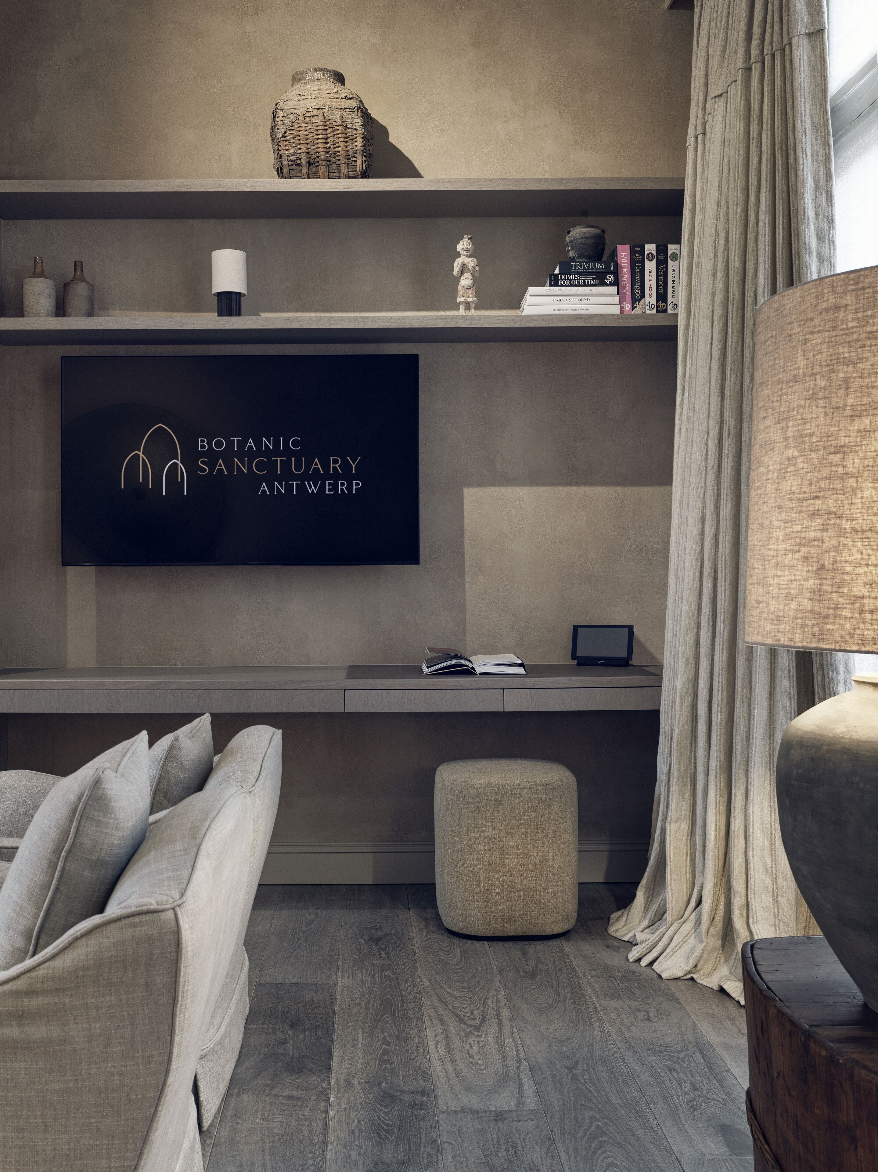 A modern living room with a grey sofa, a small ottoman, a flat-screen TV mounted on a wall, and a bookshelf with books and decorative items.