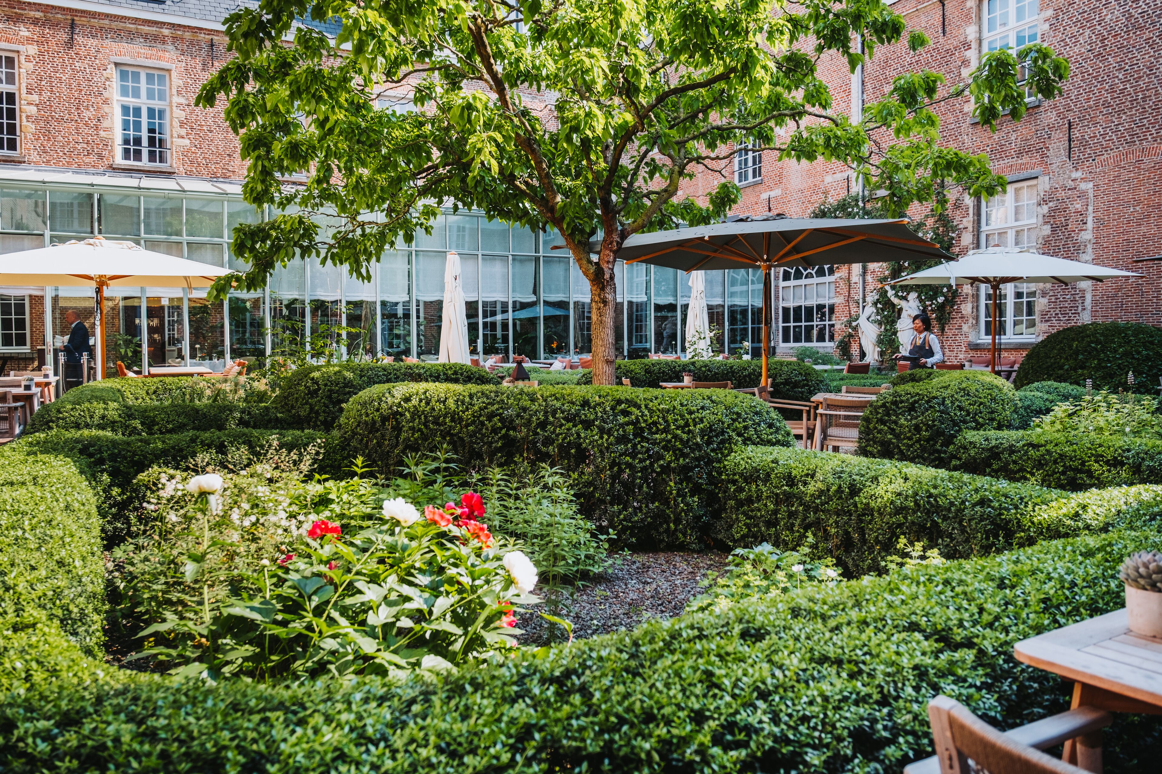 A well-maintained garden with neatly trimmed hedges, a central tree, and outdoor seating areas with umbrellas.