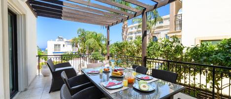 A covered outdoor dining area with wicker furniture and a glass table, surrounded by a balcony railing and palm trees.