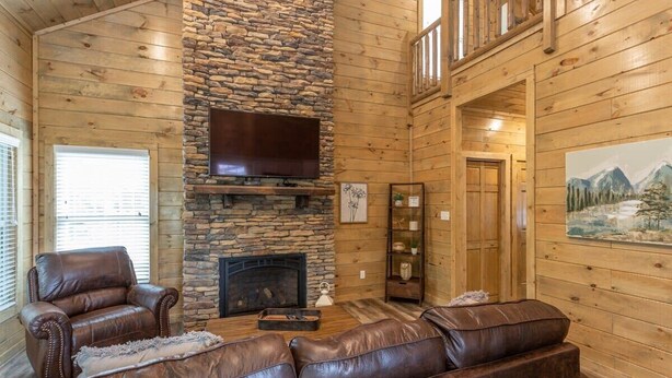 L'intérieur d'une cabane en rondins, avec une cheminée en pierre, des canapés en cuir, un téléviseur à écran plat et un tableau accroché au mur.