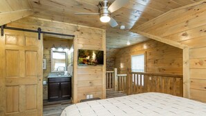 Une chambre dans un chalet en bois, comprenant un lit, un ventilateur de plafond, une salle de bain avec un lavabo et un miroir, et une fenêtre.