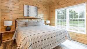 Een slaapkamer in een houten blokhut, met een bed, twee bedlampjes, een raam met zonwering en een schilderij aan de muur.