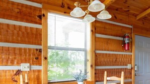 Interior de una cabaña de madera con una mesa redonda de madera y cuatro sillas, una ventana con persianas y un extintor de incendios montado en la pared.
