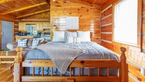 A cozy log cabin bedroom with a wooden bed, a nightstand, and a view of the kitchen.