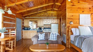 A cozy cabin interior with a wooden dining table, a sofa with checkered cushions, a bed with a gray blanket, and a kitchen area with wooden cabinets and a refrigerator.
