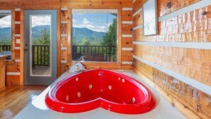 A wooden cabin with a red hot tub, large windows, and a balcony with a mountain view.