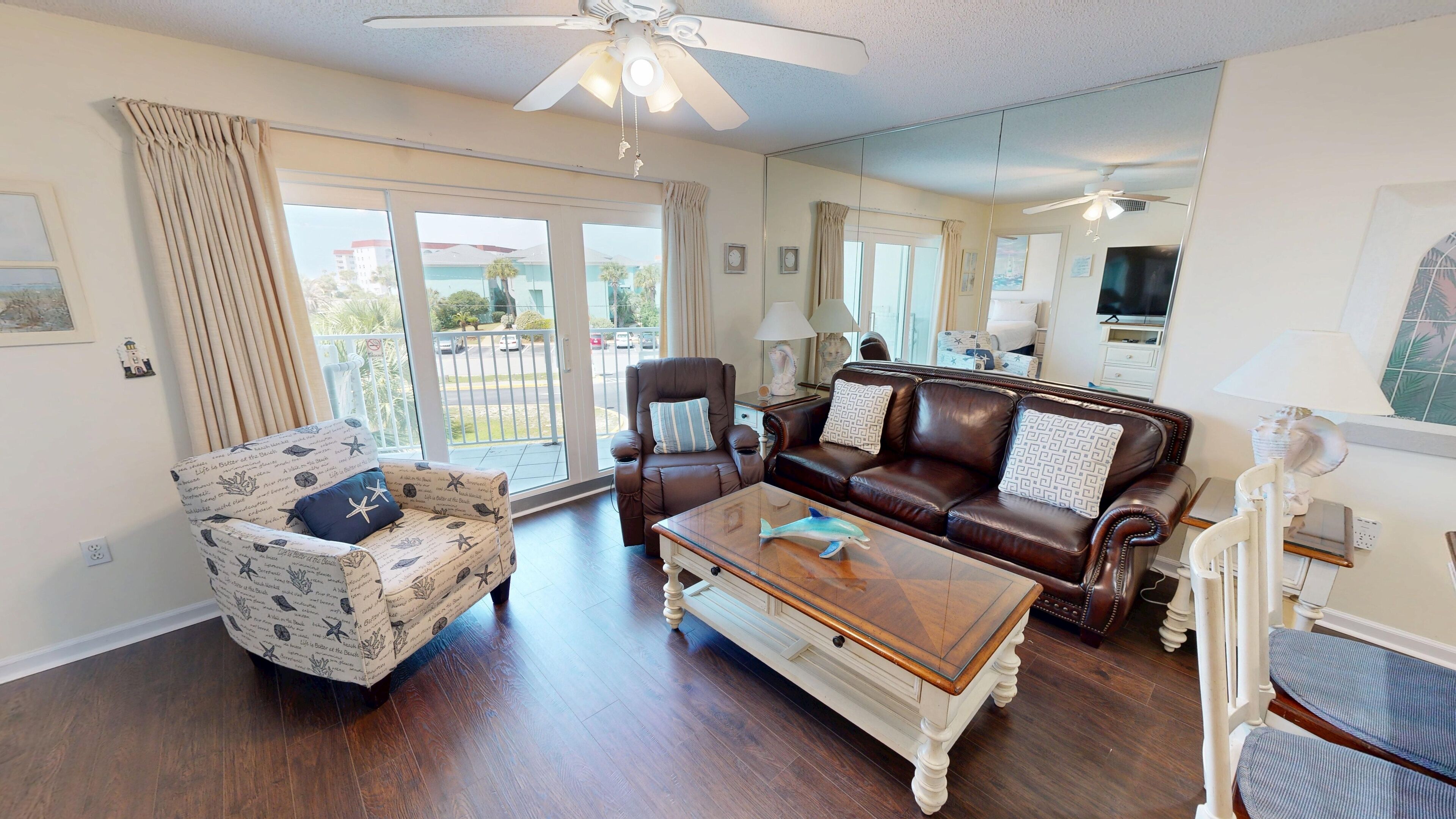 A spacious living room with a brown leather sofa, a patterned armchair, and a wooden coffee table. A ceiling fan and a large window with curtains are visible.