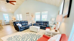Un salon spacieux avec un plafond haut, deux canapés en cuir bleu, un fauteuil rouge, une table basse blanche et un tapis à motifs corail.