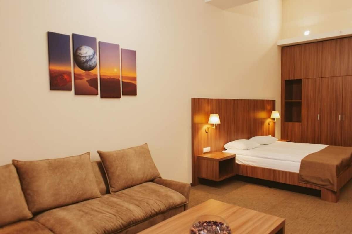 A hotel room with a bed, a sofa, a nightstand with a lamp, and wall art depicting a planet and a sunset.