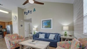 Un salon avec un canapé bleu, deux fauteuils à motifs, une table basse, un ventilateur de plafond, une horloge murale, un tableau encadré et, au fond, une cuisine.