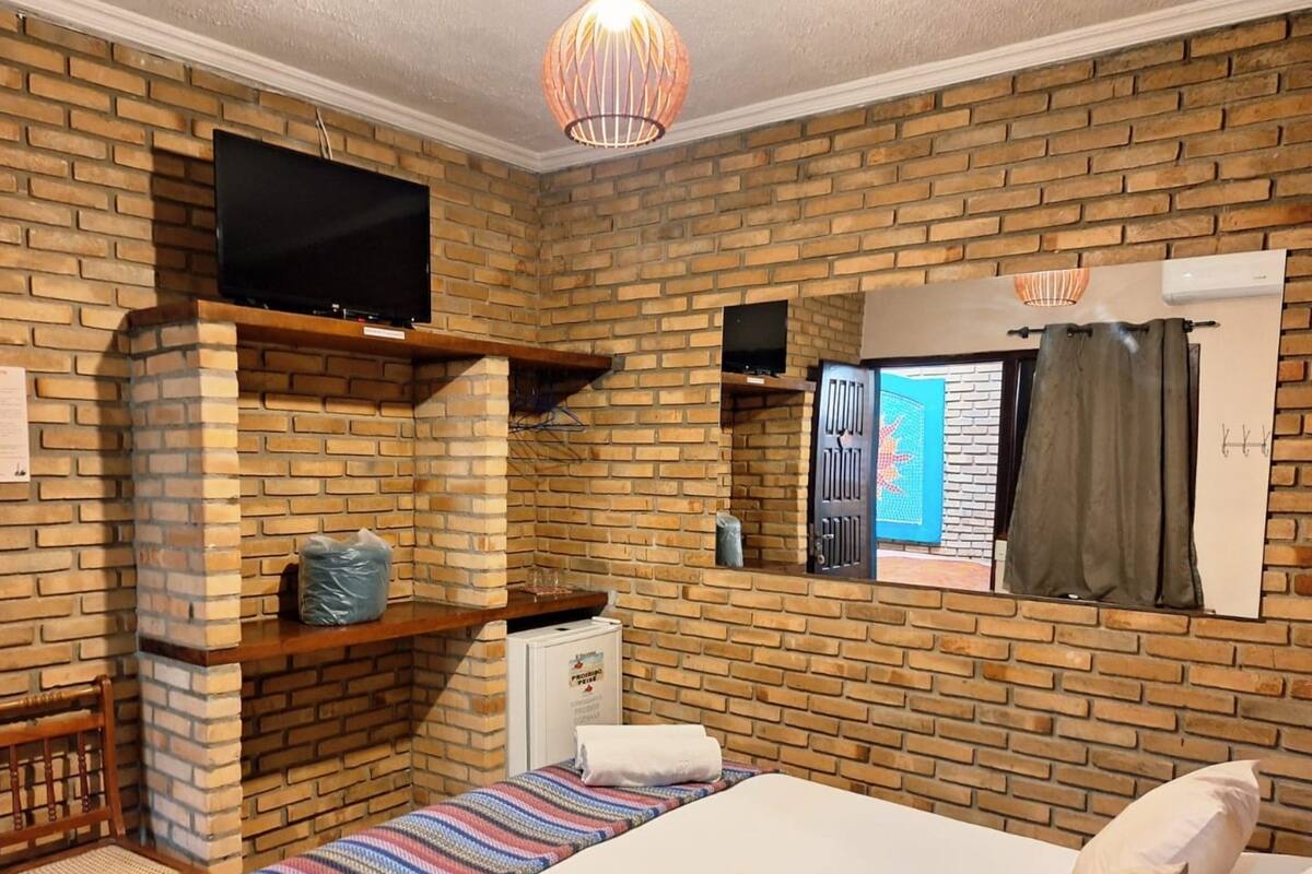 A room with a brick wall, a bed, a television mounted on a shelf, and a window with curtains.