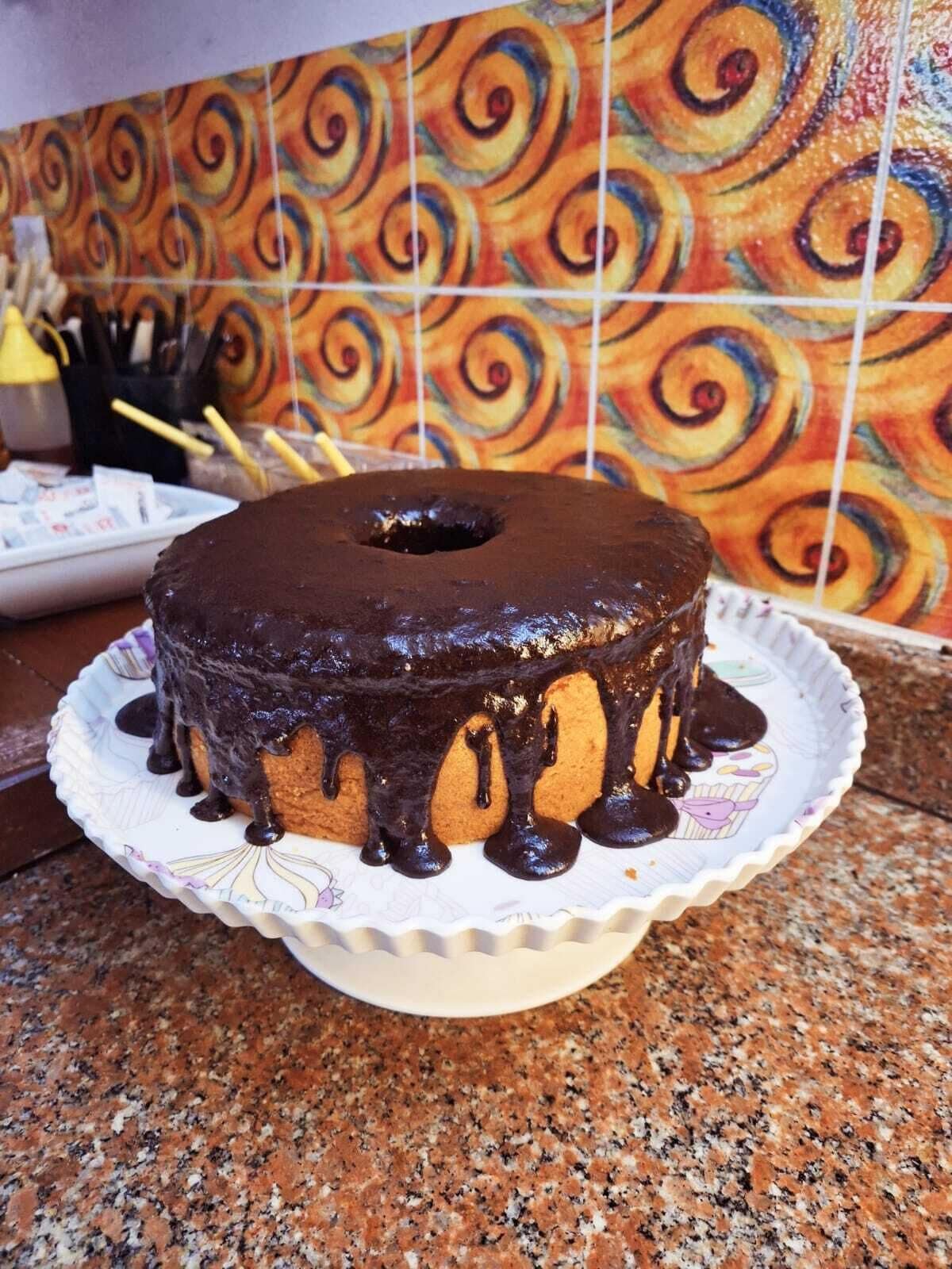 Un pastel bundt glaseado de chocolate sobre un plato de papel blanco con diseño de cupcake, sobre una encimera de granito.