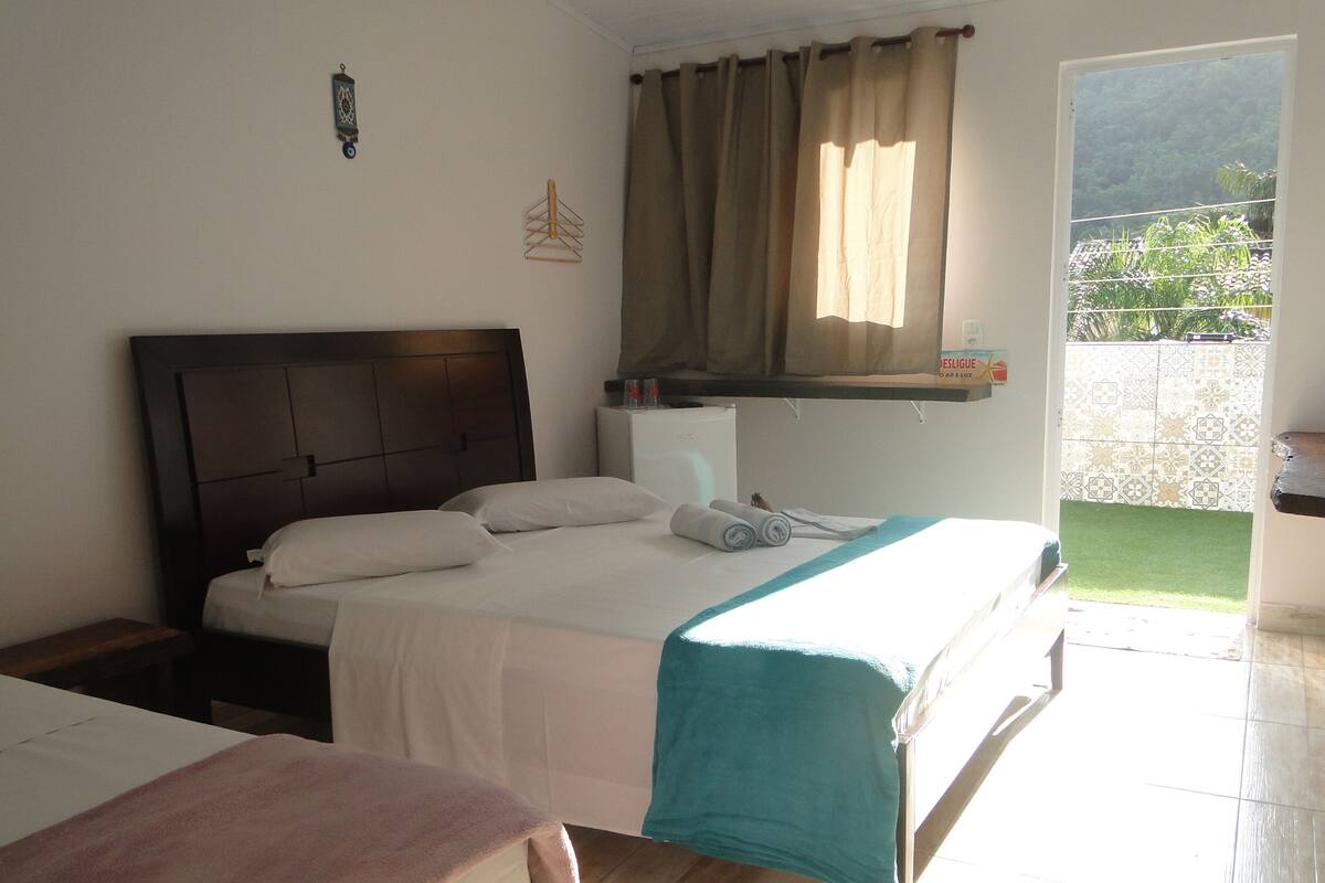 A bedroom with a bed, a nightstand, a window with curtains, a small shelf, and a view of a garden.