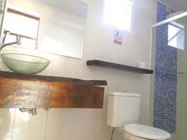 Un baño con un lavamanos de vidrio, un inodoro blanco, una zona de ducha con azulejos azules y un estante de madera.