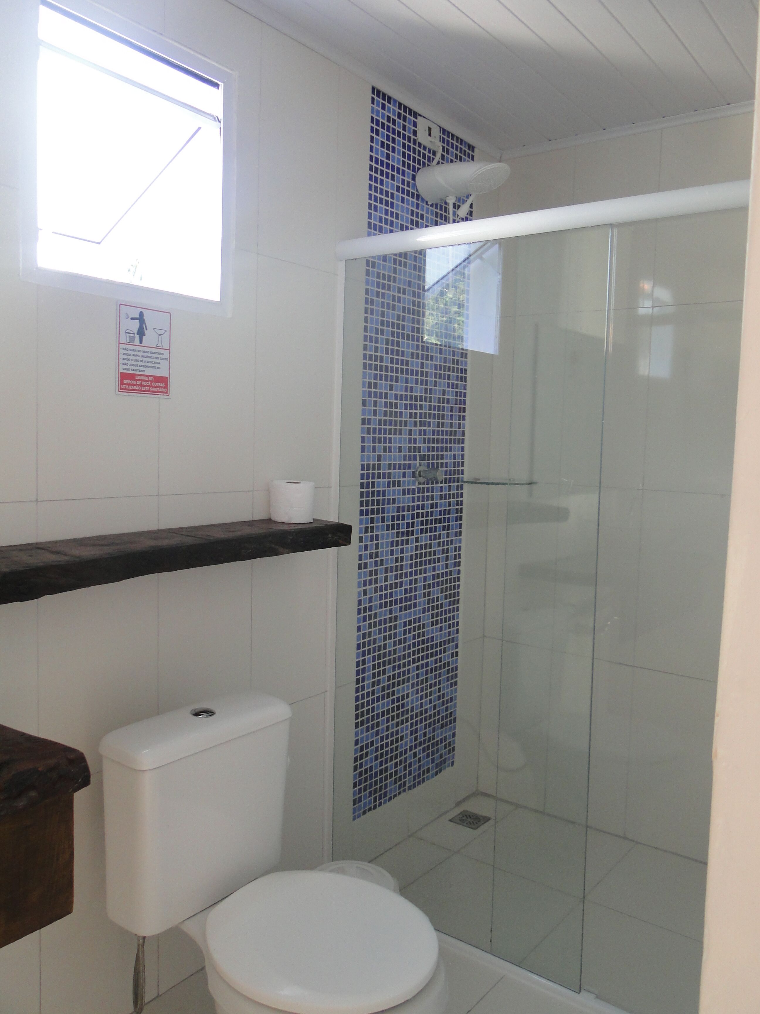 A bathroom with a toilet, a shower enclosure with a glass door, a shelf above the toilet, and a window.