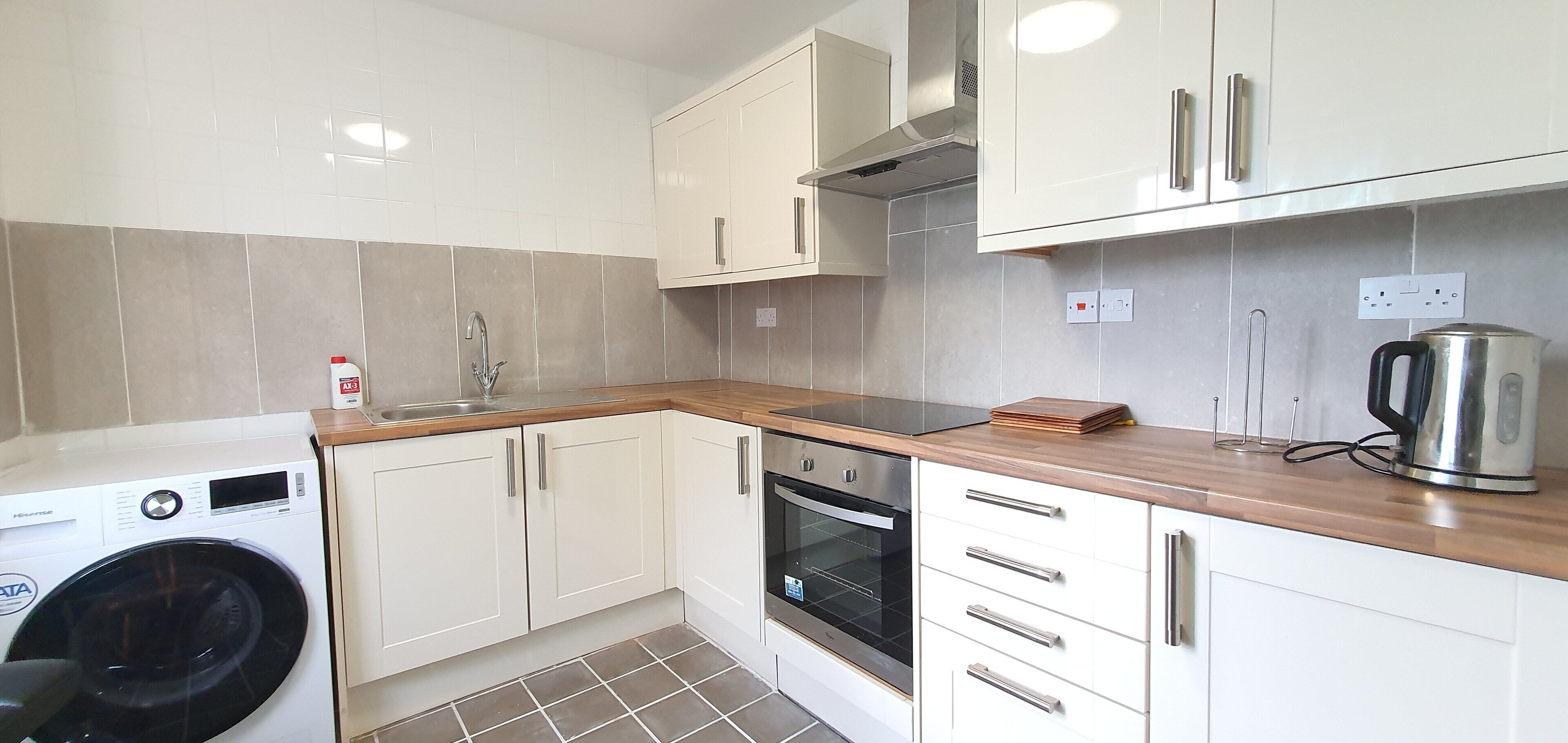 A compact kitchen with white cabinets, a built-in oven, a washing machine, and a kettle.