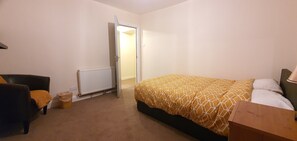 A bedroom with a bed, a chair, a radiator, and a door leading to another room. - Grace Apartments - Living Waters 1 (Ashford)