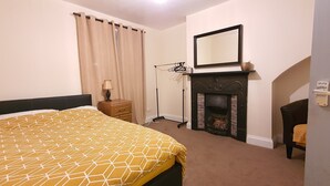 Een slaapkamer met een bed, nachtkastje, lamp, open haard en spiegel.