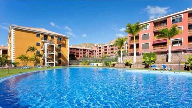 A swimming pool surrounded by multi-story buildings with balconies, palm trees, and lounge chairs. - Luxury apartment in Residencial El Mocan (Palm-Mar)