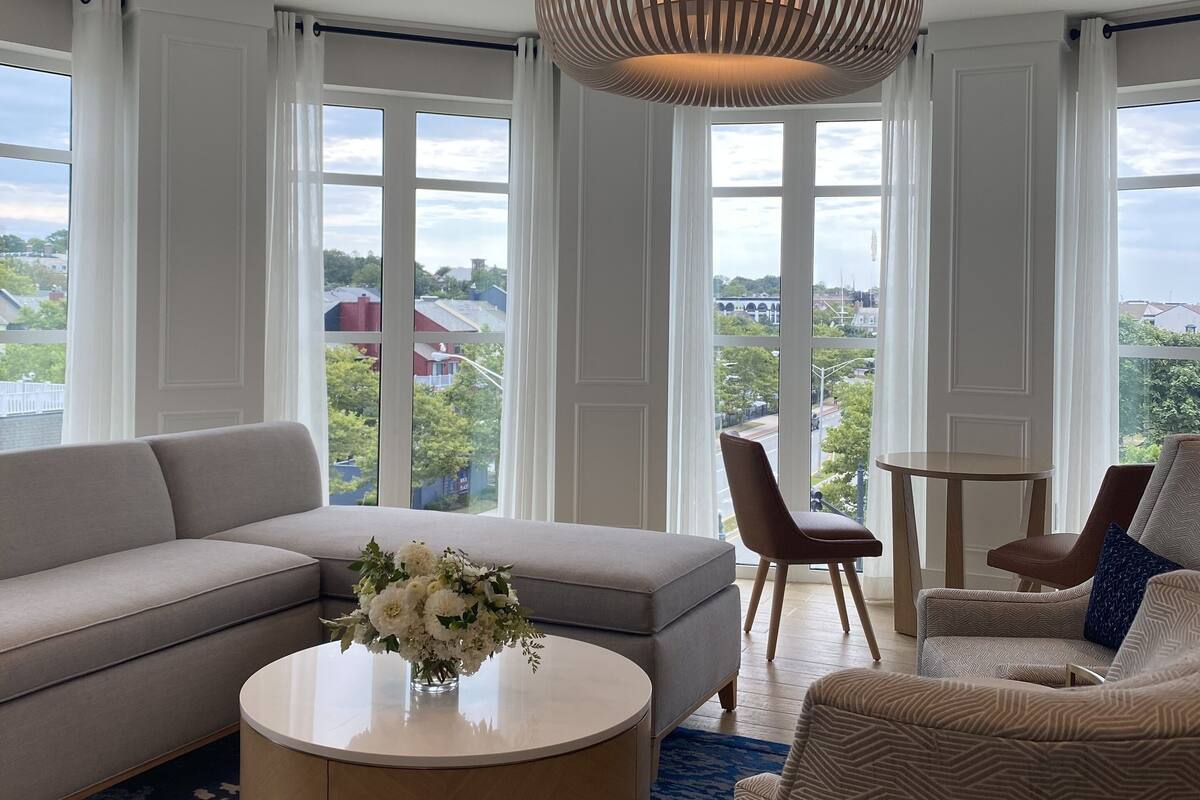 A modern living room with a large sectional sofa, a round coffee table, and a patterned armchair. There is a large window with a view of the cityscape.