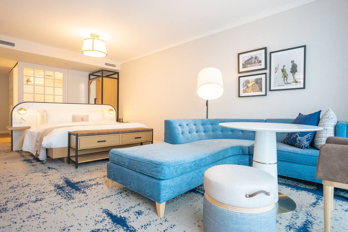 A modern hotel room with a bed, a sofa, a round table, and a small ottoman. There are framed pictures on the wall and a lamp on the floor.
