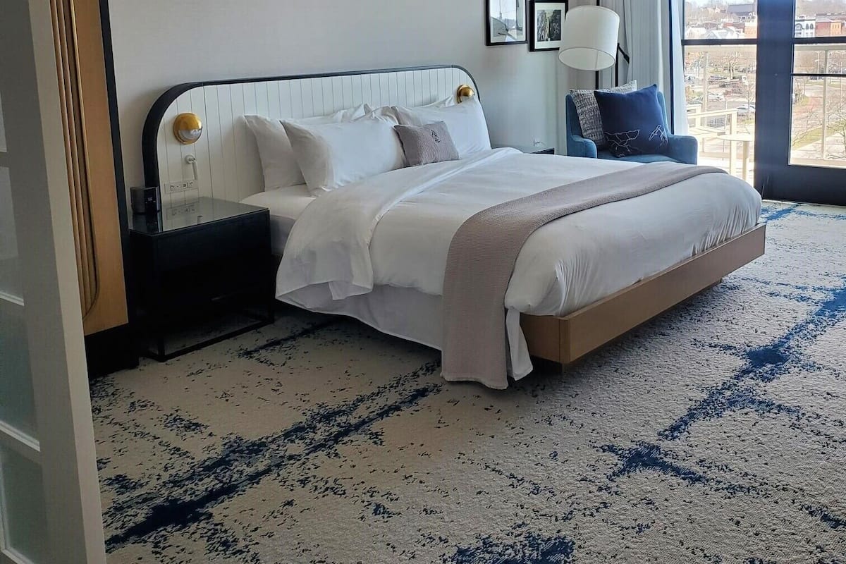 A hotel room with a large bed, a nightstand, a lamp, and a view of the city through large windows.