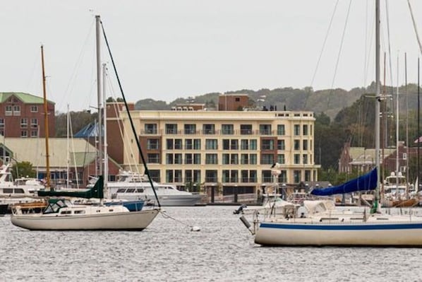 A marina with several sailboats docked, a building with multiple windows in the background, and a hilly landscape.