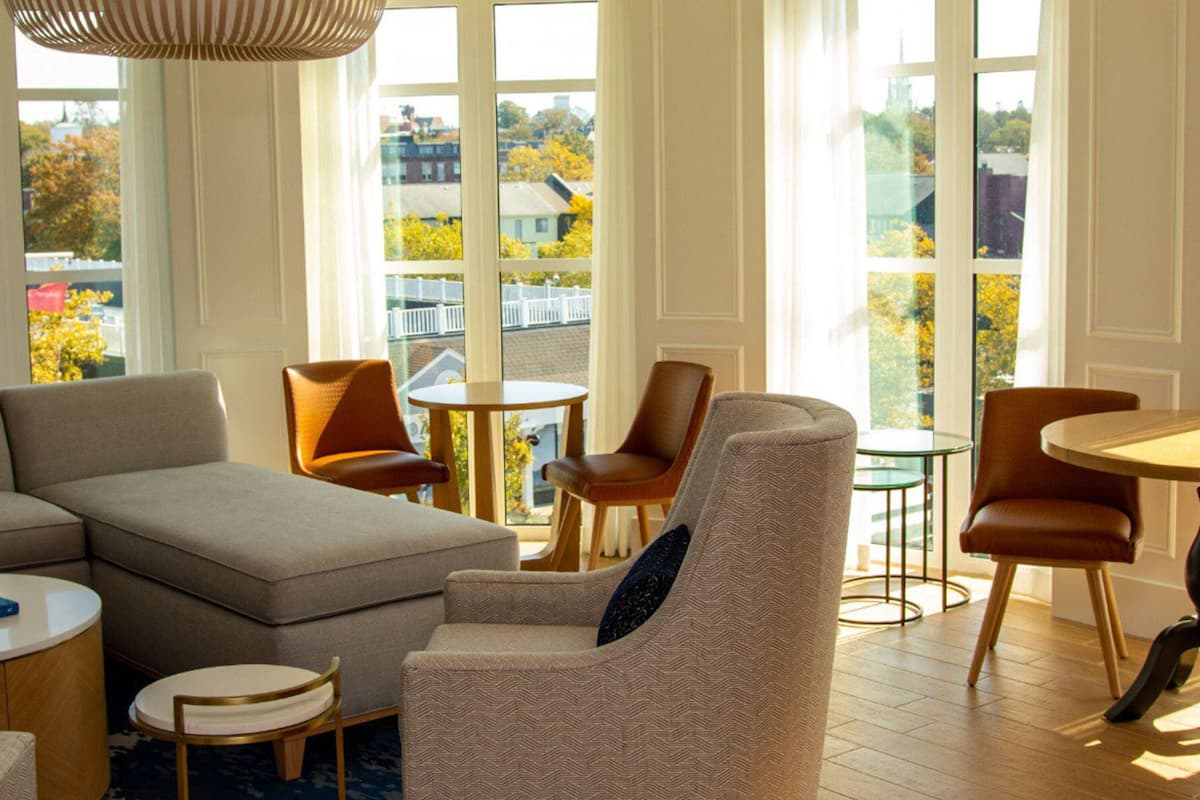 A modern living room with a sectional sofa, a round coffee table, and two armchairs. Large windows provide a view of the city.