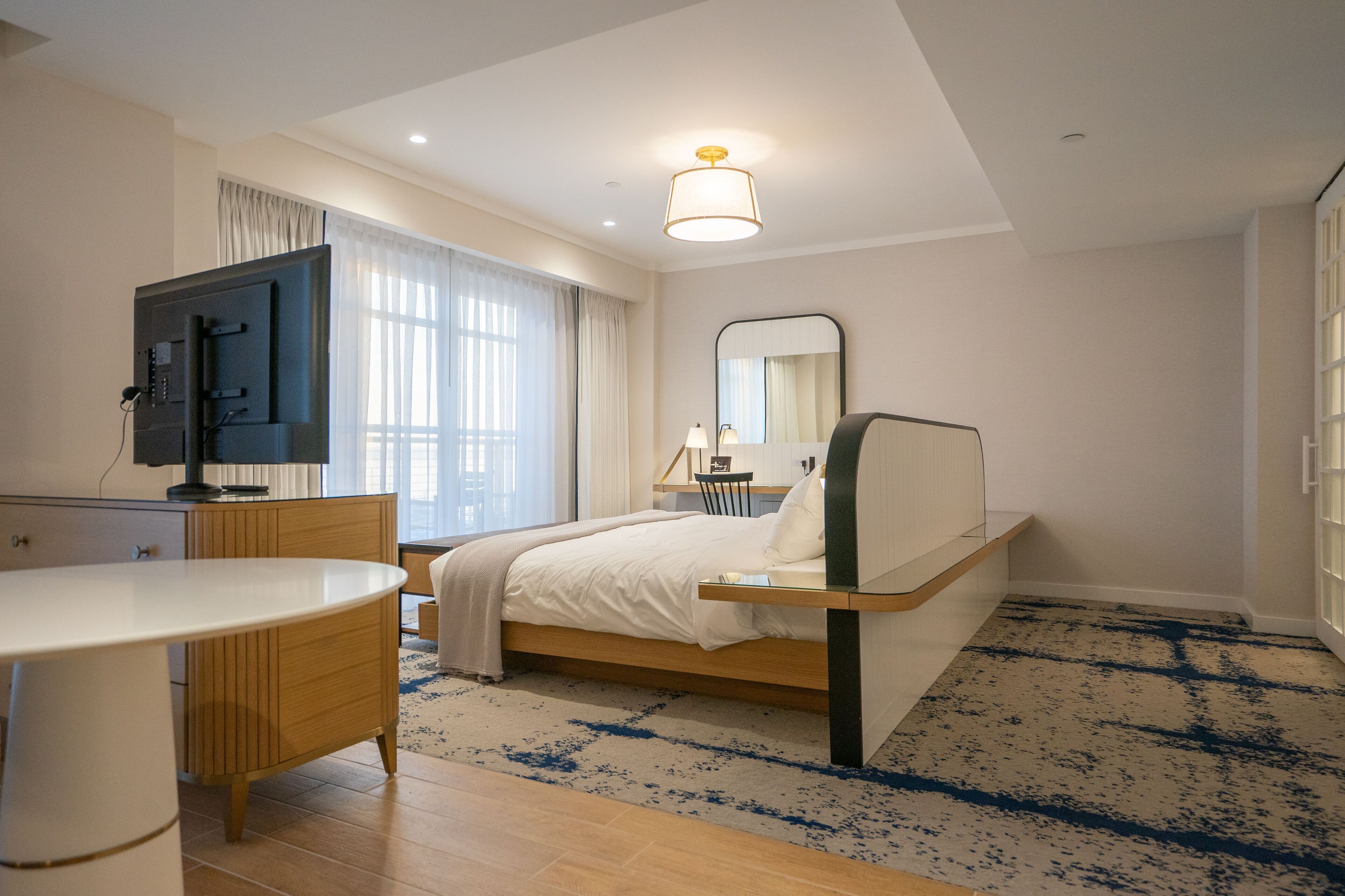 A modern hotel room with a bed, bedside table, TV, and a large window with curtains.