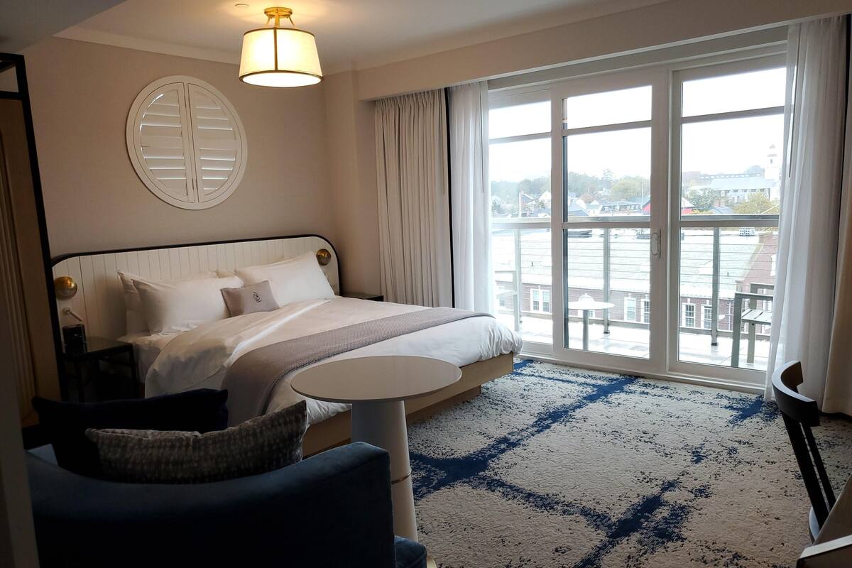 A hotel room with a large bed, a round table, and a view of a body of water through large windows.