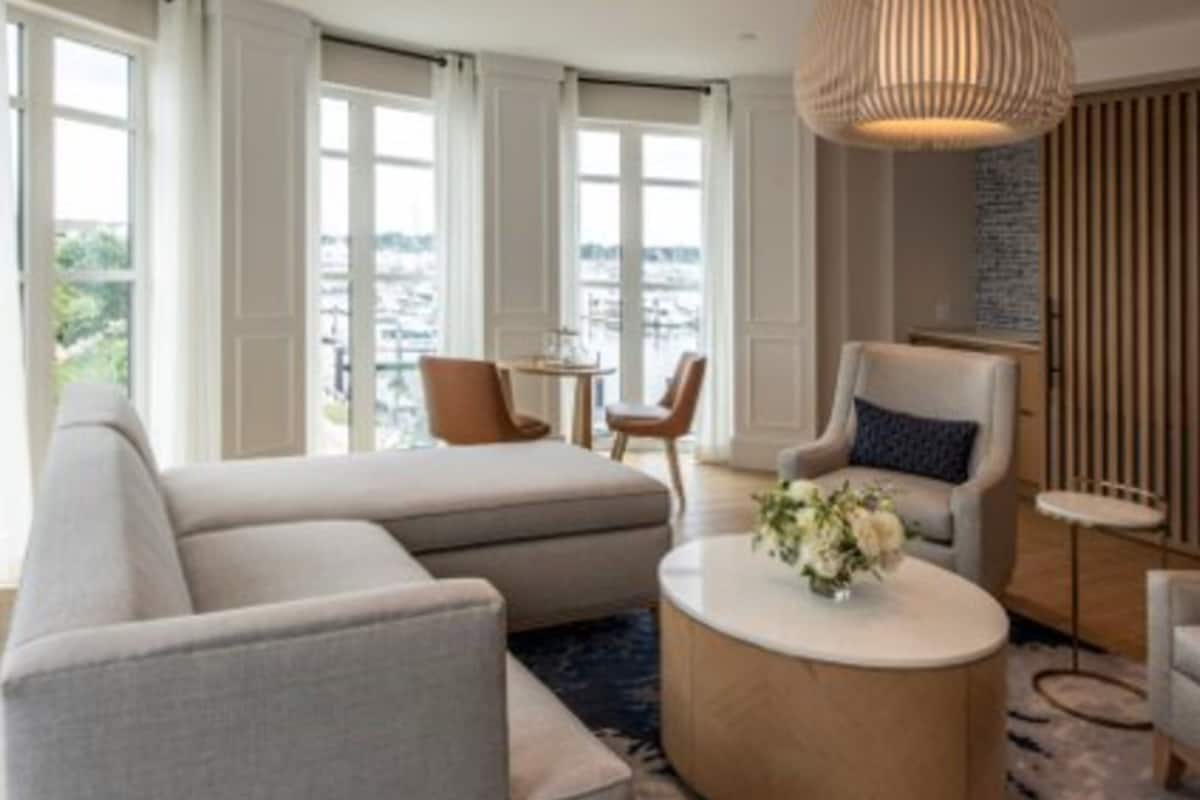 A modern hotel room with a large sectional sofa, a round coffee table with a vase of flowers, a comfortable armchair, and a view of the city through large windows.